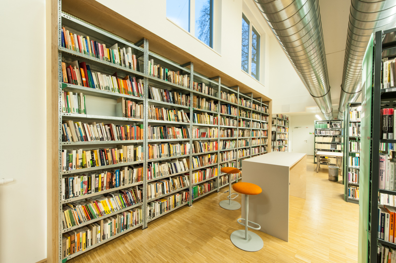 Zu sehen ist ein langes und hohes Bücheregal in der C3 Bibliothek. Davor steht ein Schreibtisch mit zwei Hochstühlen