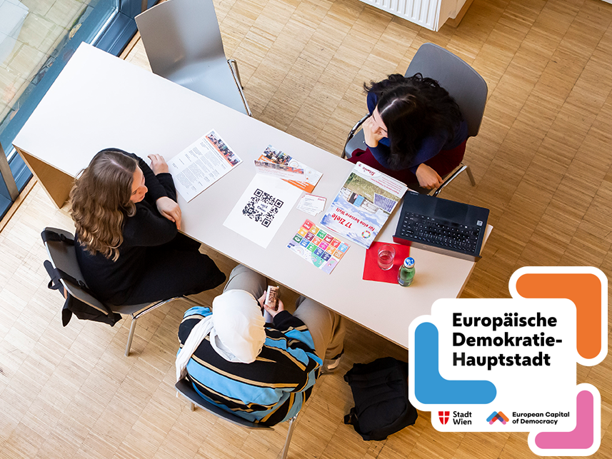 Drei Personen im Gespräch in der C3-Bibliothek von oben aufgenommen, Logo der Stadt Wien: Europäische Demokratiehauptstadt